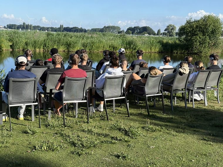 Eine Schulgruppe sitzt mit Kopfhörern in Reihen am Wasser