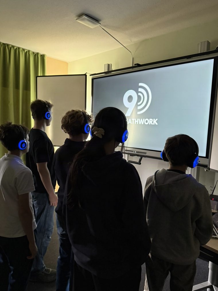 Schüler stehen mit Kopfhörern vor dem Board, auf dem das 9D-Zeichen läuft